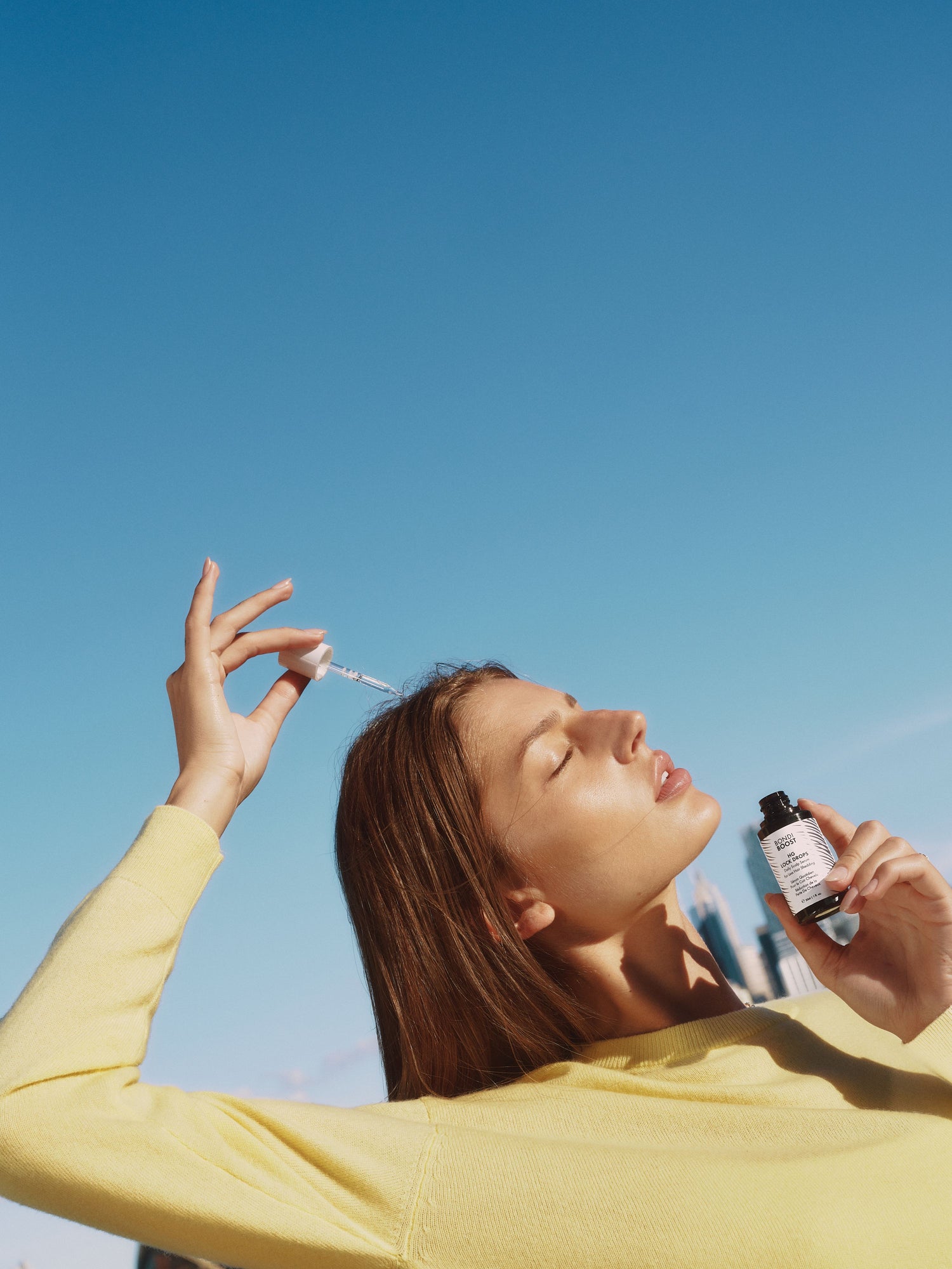 Woman applying a dropper of skincare product against a clear blue sky